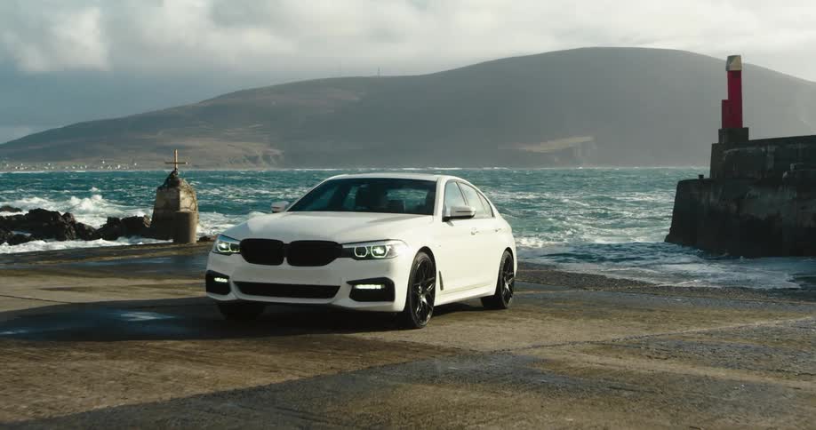 A white car is parked on pavement next to an ocean with a hill on the opposite side