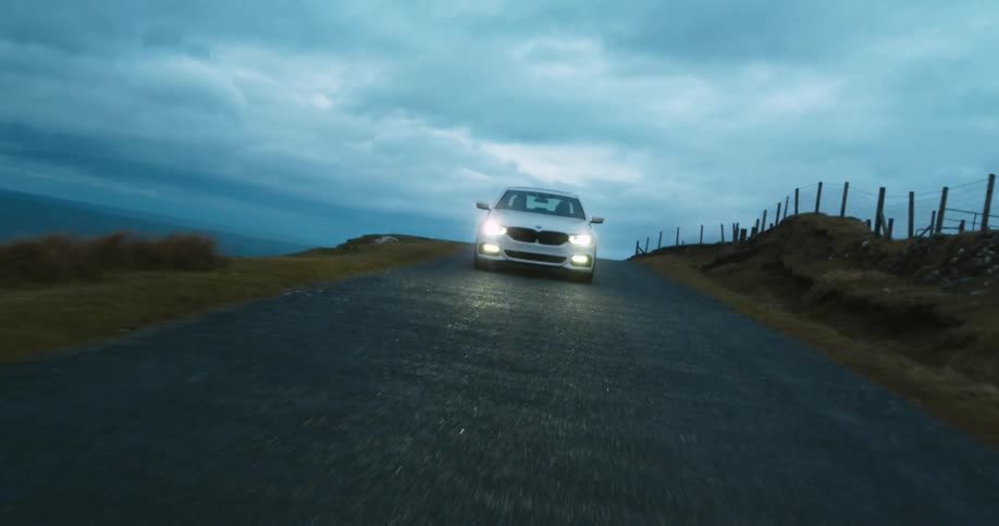 Overcast sky at dusk to a white car driving with headlights on down a remote paved road