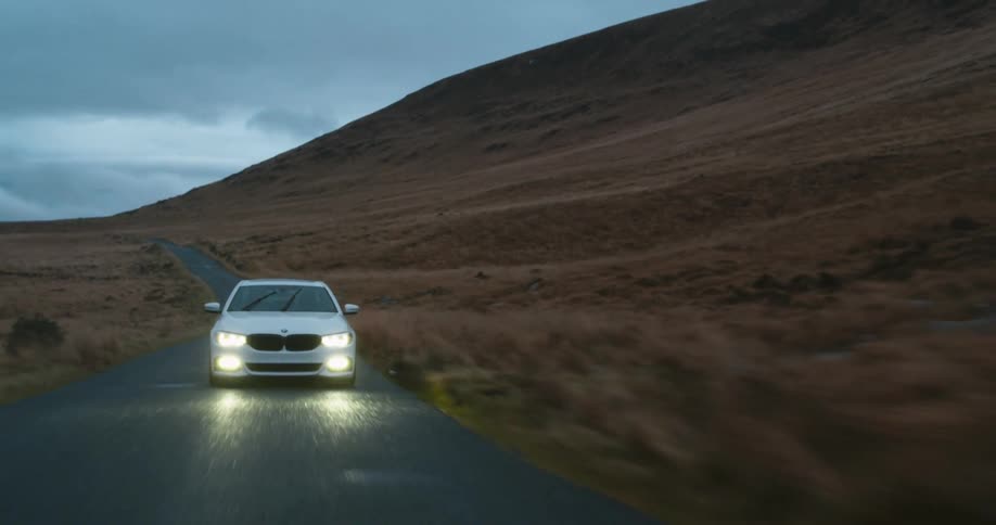 White car driving on road next to mountain with headlights on