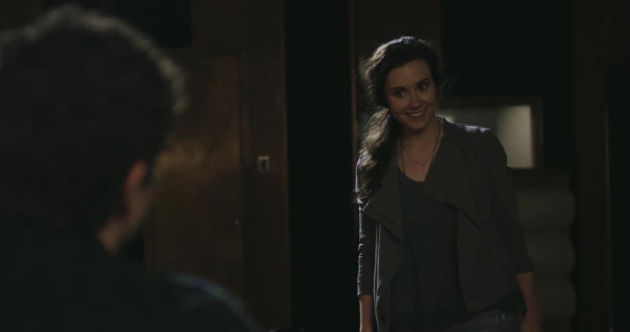 A woman carrying a guitar takes approaches and speaks to a man in a dimly lit room