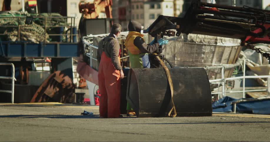 Two men stand in front of a machine while one of them hits one of its parts with his hand at daytime