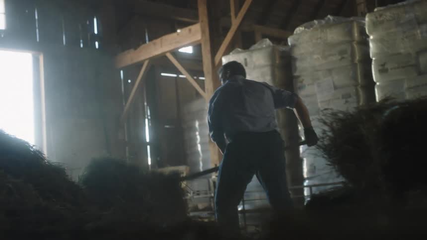 A man inside a dimly lit barn moving hay using a fork in a ranch