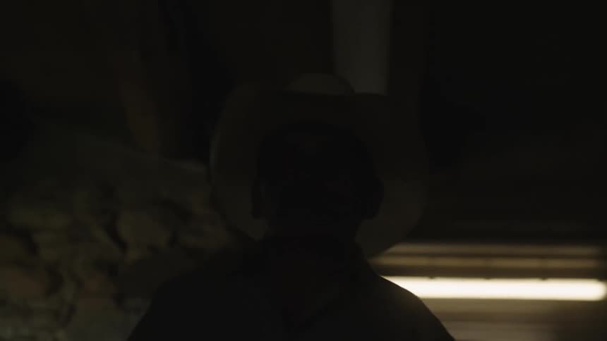A man wearing a cowboy hat walks through a stable with overhead lighting while carrying reins