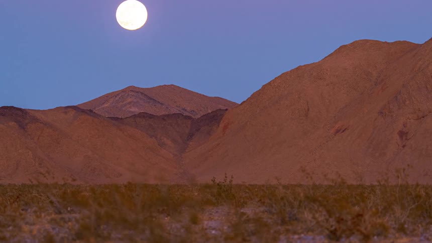 The moon rises over the mountains in a blue and pink sky
