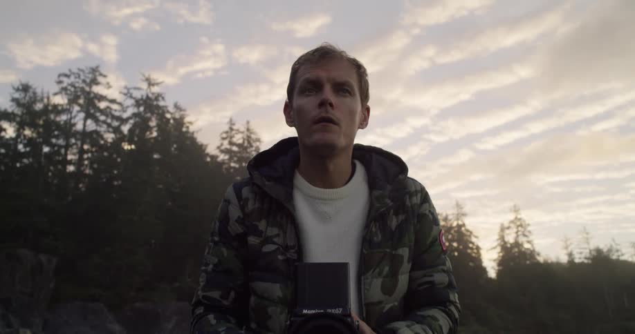 A man takes a film photo on the beach at dawn with forest and clouds