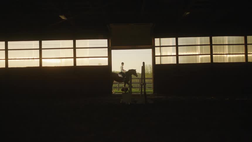 A couple horses with riders passing in front of a stable door at sunset