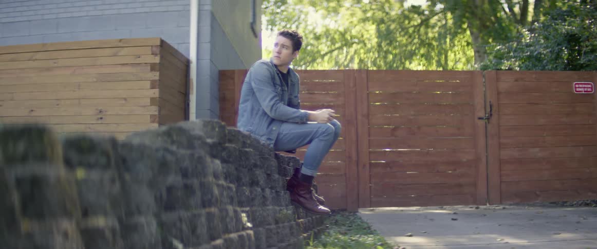 A young man sits on a wall of stacked stone bricks in front of a house next to a gated road