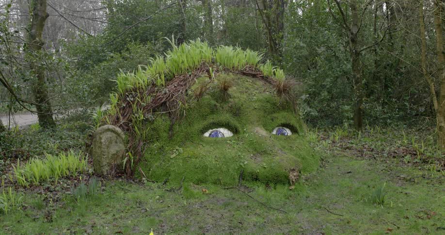 Rain falling over a sculpture of a buried head covered in moss and grass in a forest