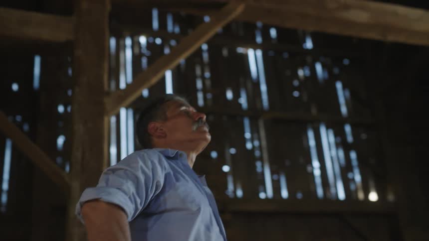 A man looking up stands inside of a run down wooden barn where wall boarding is missing