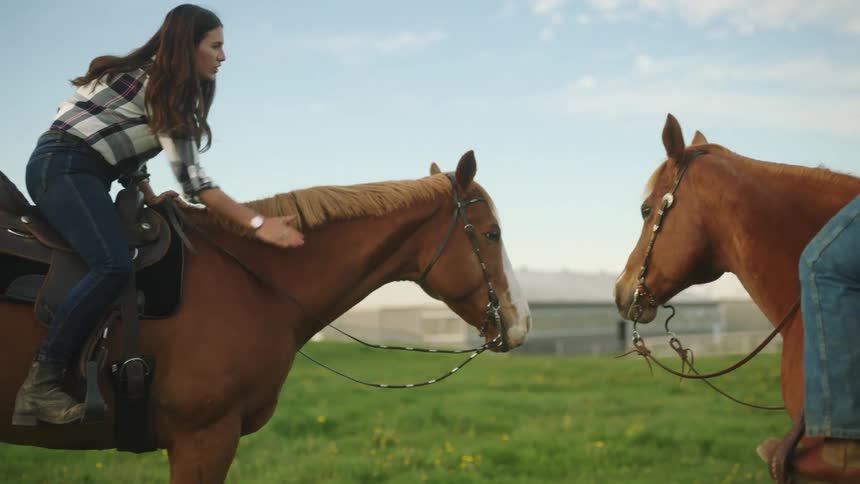 A woman sitting on a horse in a pasture with a man on horse pats her horse on the neck