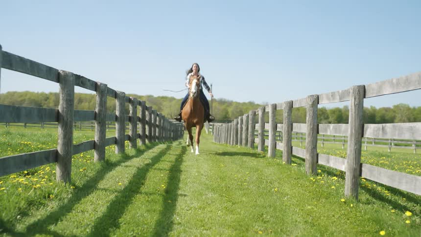 Young woman riding a horse between wooden fences in the green grass of a ranch