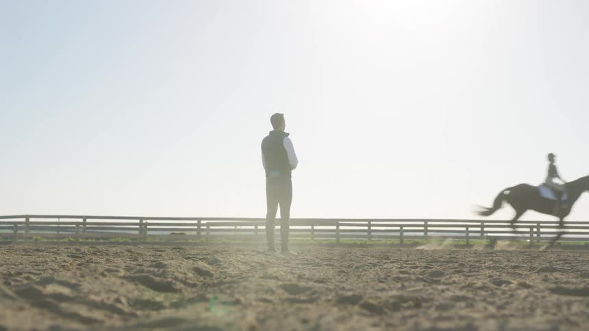 A man stands in a pasture and watches a girl riding a horse