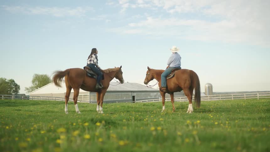 Man and woman riding two brown horses in the middle of a grass field at a farm