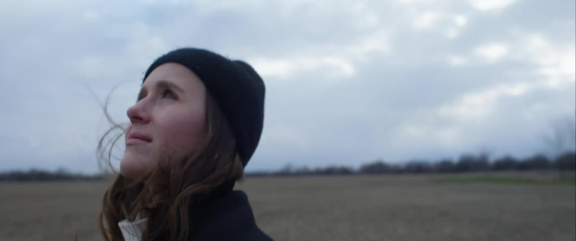 A woman stands on a field with brown grass and overcast skies wearing a knot hat and coat