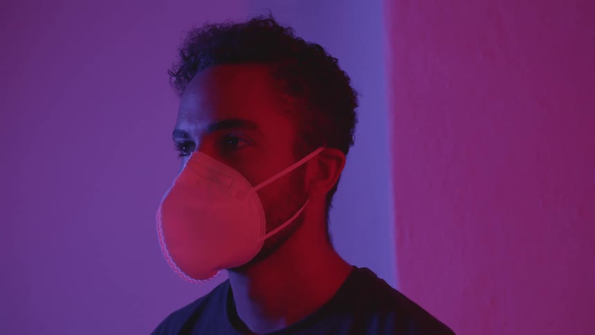 Young man puts a face mask on while standing in a white room under red and purple lights
