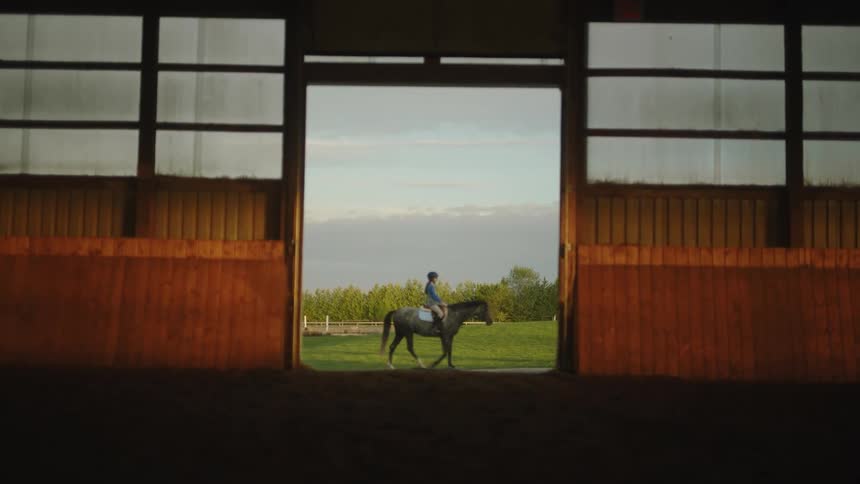 A woman rides a horse past the opening of a stable during the day outdoors