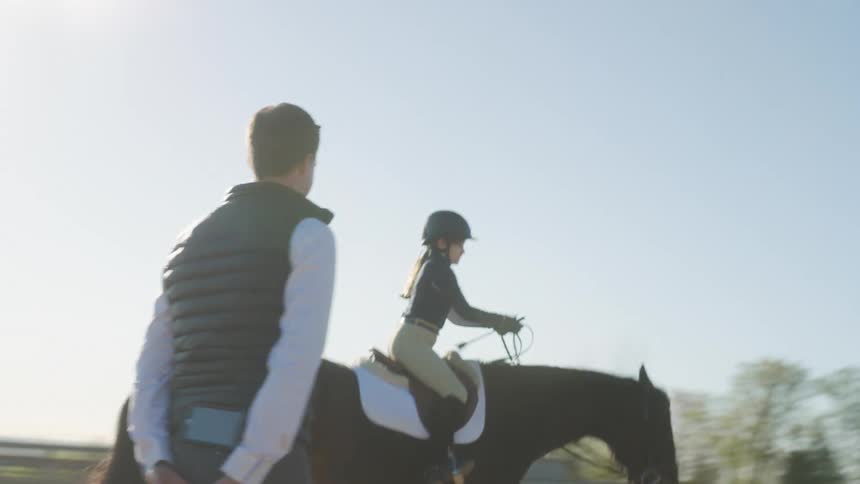 A girl rides a horse outdoors with a man coaching her