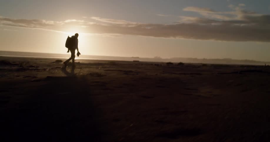 A silhouetted person wearing a backpack walks along the beach at sunset