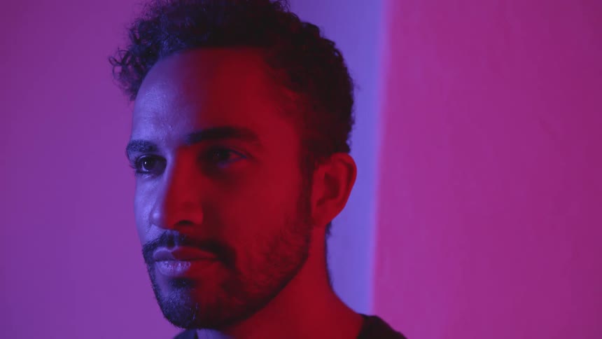 A man stands against a pink lighted corner of a wall while opening and closing his eyes