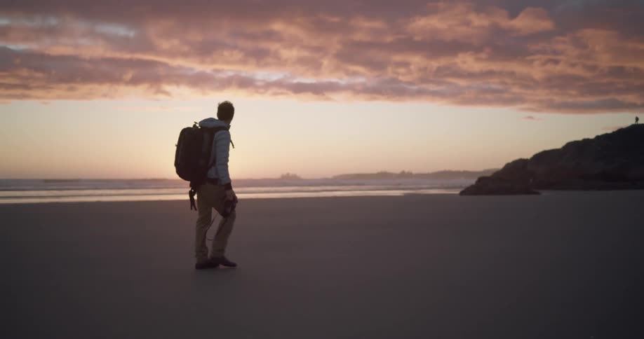 Man wearing backpack walks across beach as setting sun turns cloud cover and sky red