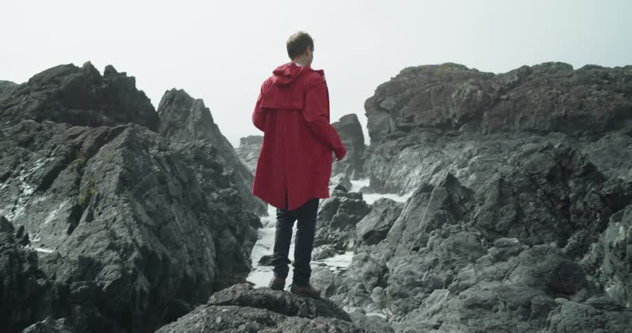 Man wearing bright red coat standing over the jagged rocks on a beach