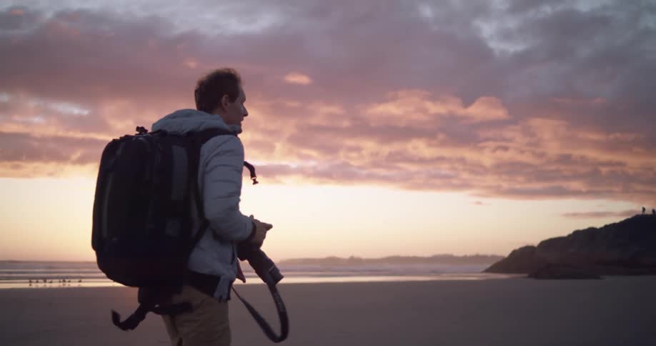 A photographer is carrying a large camera on the beach at sunset