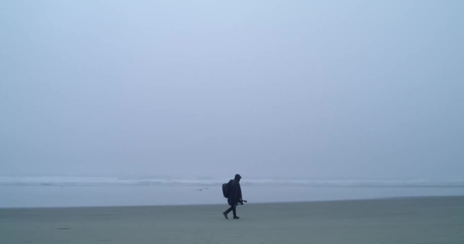 A person walks along a beach in misty overcast conditions wearing an overcoat and carrying a backpack