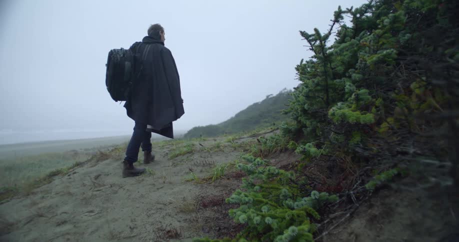 A man wearing a coat and carrying a backpack hiking up a sandy hill from beach