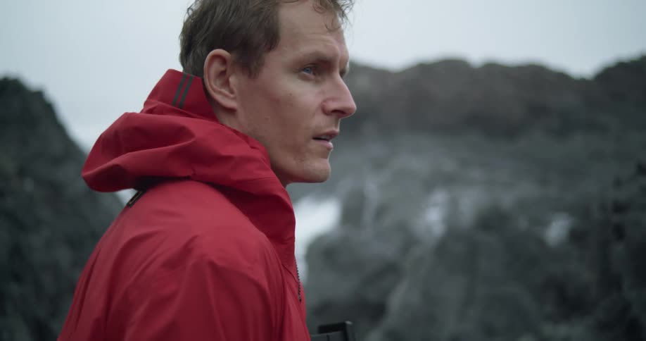 A man in red jacket holding camera turns towards ocean rocks