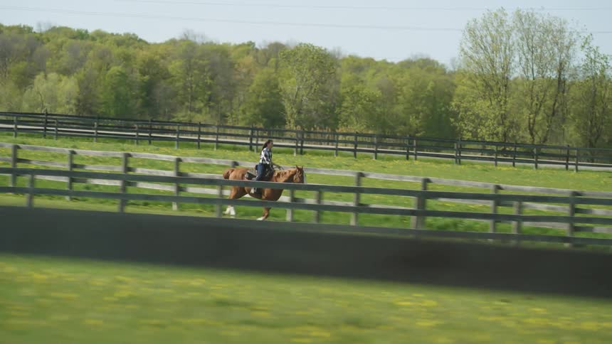 A woman rides a horse between large horse corrals in the countryside during the day