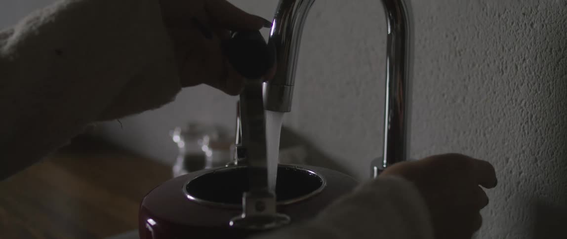 A woman holding a silver tea kettle under water coming from a kitchen faucet turns off water and moves the kettle away