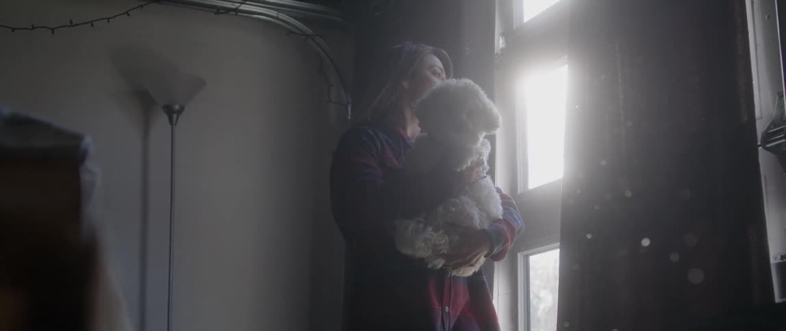A woman holds her white dog as they look out a window of her apartment
