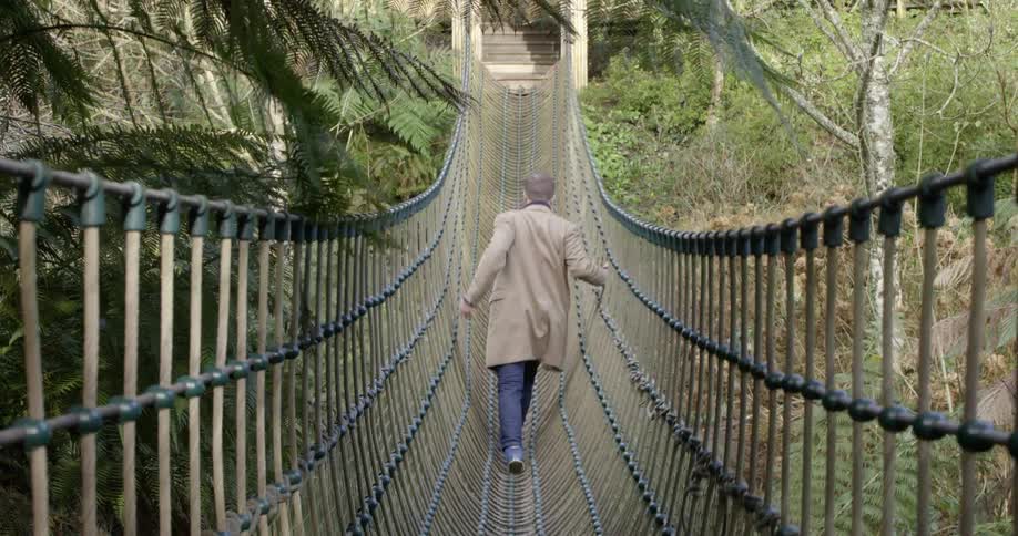 A man in a long camel coat walks across a narrow rope suspension bridge