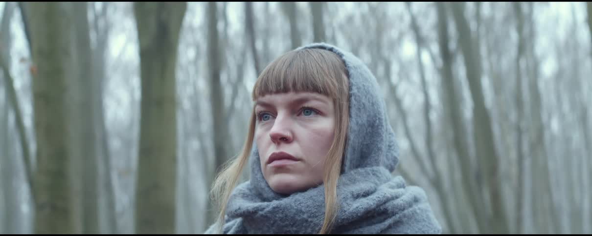 A woman with a scarf standing in a forest with trees the sky and clouds in the background