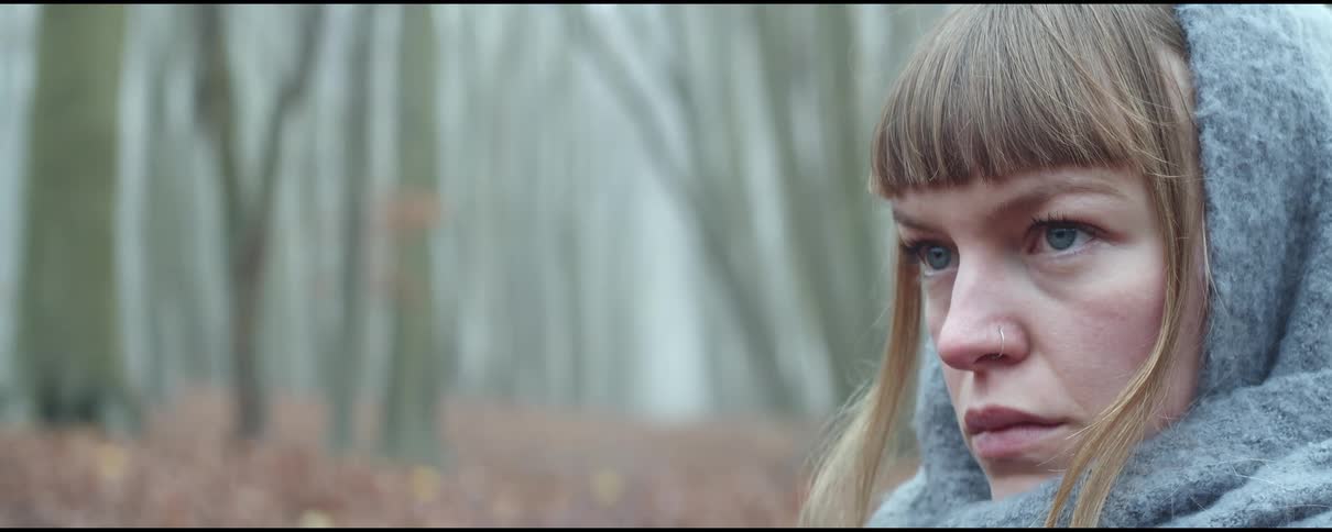 A woman with a scarf over her head stands in a forest and looks off in to the distance