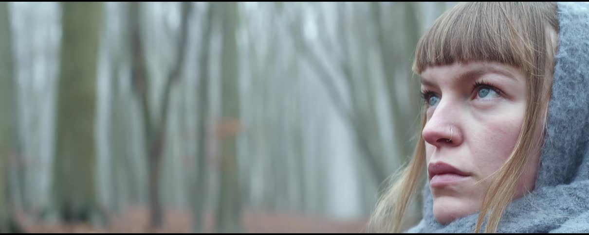 A young woman with her head and neck wrapped in a scarf stands in a forest devoid of leaves during the day