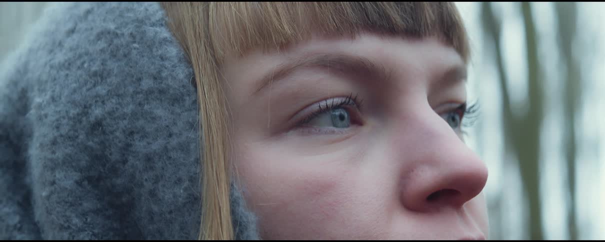A young woman in cold weather clothing stands in a woodland during the day