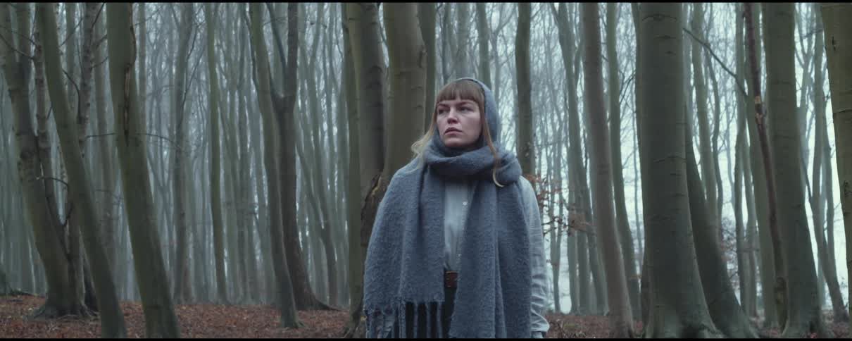 A woman stands in a forest wearing a large knit scarf