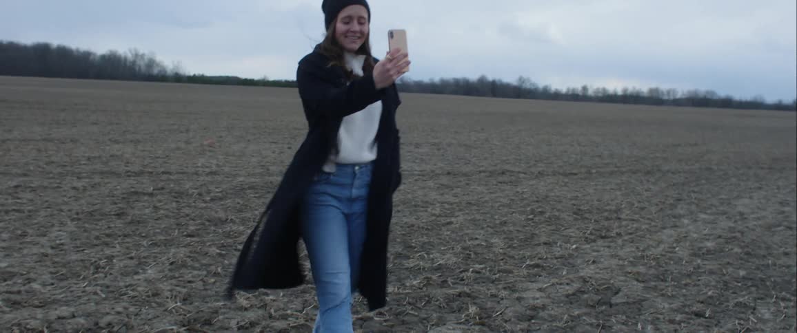 A young woman in coat weather clothing uses her smartphone to film herself as she walks through a field on a cloudy day