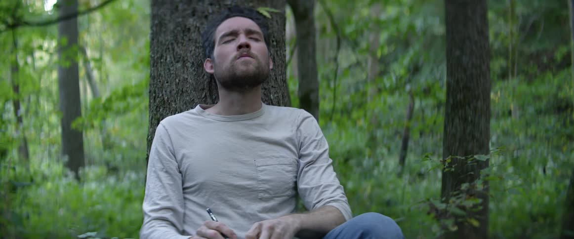 A man with a beard sits against a tree in a forest writing on a pad