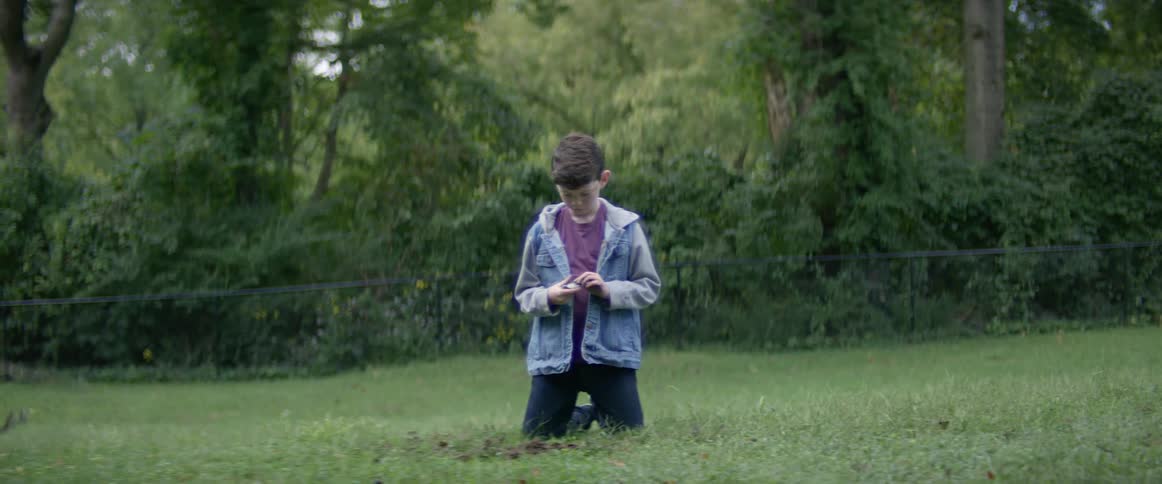 Boy wearing jacket kneels in grass in mowed field examining item