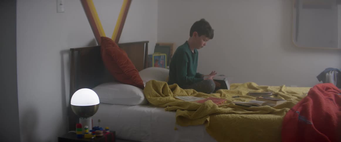 A boy looks through a book while sitting on a bed covered in books in his bedroom