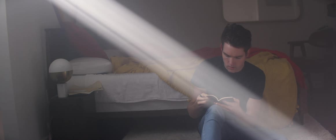 A teenage boy sitting on the floor at home picks up a notebook and writes in it