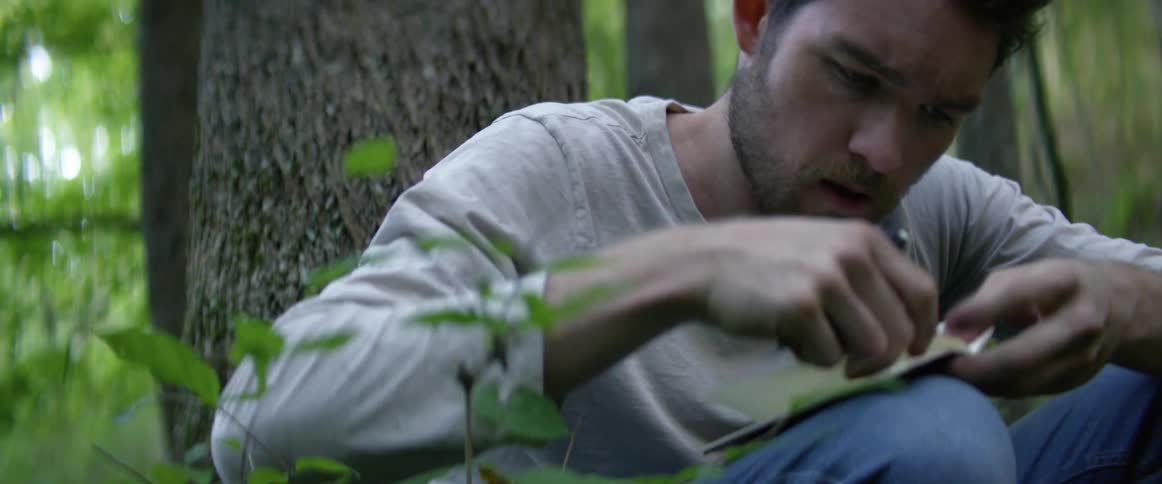 A man sitting in a forest writing in a book then ripping a page out