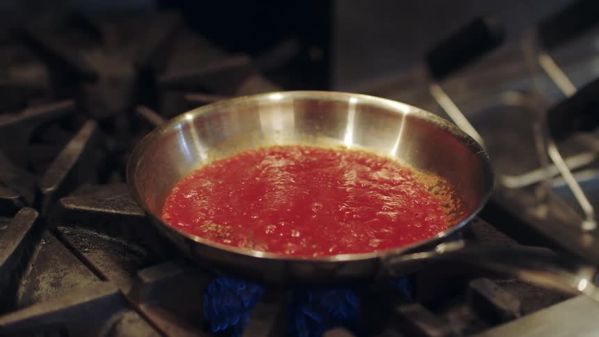 A skillet with red sauce bubbles on a stove top heated with a gas flame