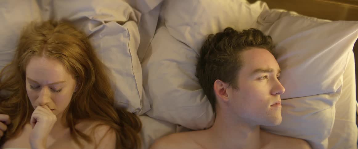 A woman lying next to a man in bed looks at him and smiles and he turns his away and then she frowns and bites her finger