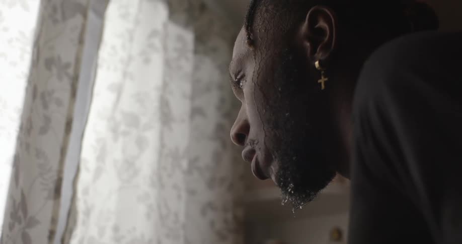 A black man stands at a window indoors with water dripping from his goatee