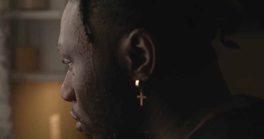 A man with a cross earring in his ear and beats of sweat sliding down his head indoors