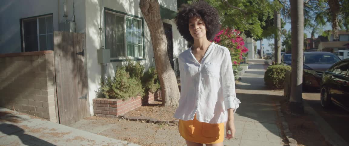 Brunette woman with curly hair white shirt and orange mini skirt walks down the street