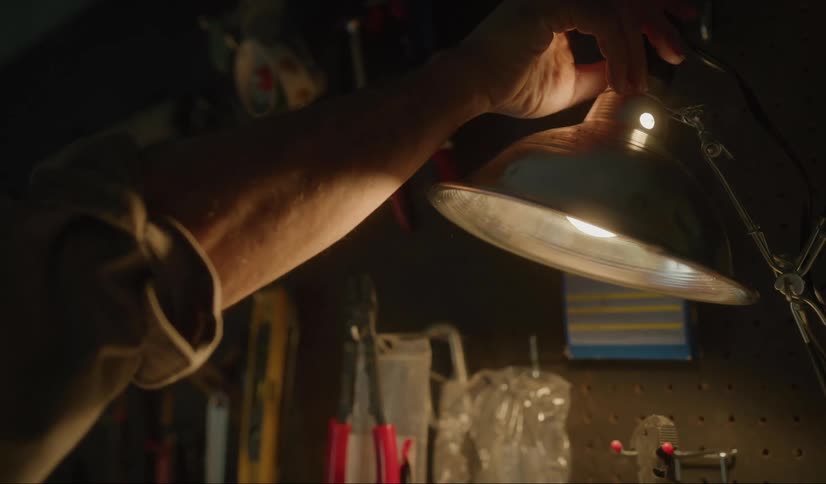 A man reaches over to the top of a lamp in a dark workshop and turns the light on